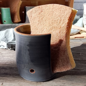 Könnte beinhalten: Eine dunkelgraue Keramik-Schale mit einem Loch an der Seite, die einen natürlichen Schwamm hält. Der Schwamm besteht aus einem hellbraunen Material mit einer faserigen Textur.