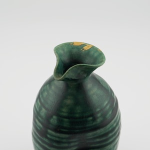 May include: A green ceramic bottle with a wide, flared opening and gold accents. The bottle has a textured surface and a rounded shape.