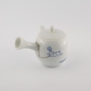 May include: White ceramic teapot with a lid and a spout. The teapot features a blue illustration of a running rabbit. The handle has a gold repair. A small, round lid sits on top. The teapot is designed for brewing tea.