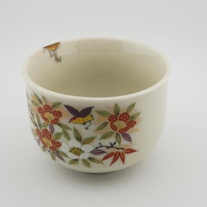 May include: A white ceramic teacup with a floral design. The design features red, yellow, and white flowers, green leaves, and purple and blue birds. The cup is decorated with gold accents.
