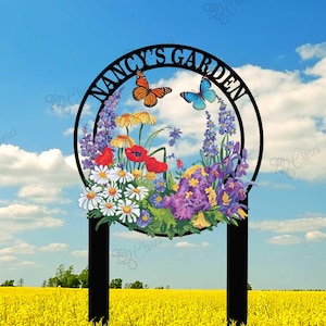 May include: Black metal garden sign with a circle frame and the text "NANCY'S GARDEN". The circle frame has a floral design with butterflies and a variety of flowers in shades of pink, purple, red, yellow, and white. The sign is standing in a field of yellow flowers.