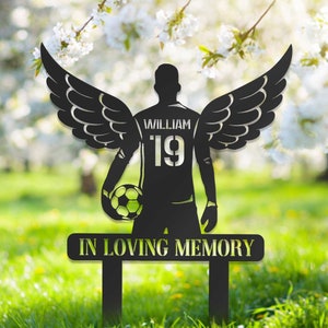 Könnte beinhalten: Schwarze Metallsilhouette eines Fußballspielers mit Flügeln, der einen Fußball hält. Der Spieler trägt ein Trikot mit dem Namen "William" und der Nummer "19". Das Schild lautet "In liebevoller Erinnerung".