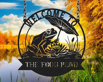 Może przedstawiać: Czarny metalowy znak powitalny z żabą trzymającą parasol nad liściem lilii wodnej. Znak głosi "Witamy w The Frog Pond".