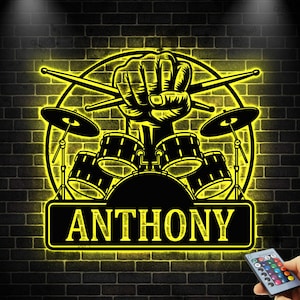 May include: A black and yellow illuminated drum kit sign with the name "ANTHONY" displayed on a black brick wall. The sign features a fist holding drumsticks, a drum kit, and cymbals.
