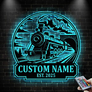 May include: A black and blue neon sign with a train design. The sign is circular, showing a train, tunnel, and tracks. The text "CUSTOM NAME" and "EST. 2025" are displayed on a rectangular banner. A remote control is visible in the bottom right.