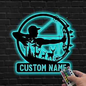 Op de afbeelding: Zwart metalen bord met een silhouet van een boogschutter die een pijl en boog afvuurt, omgeven door een blauwe gloeiende cirkel. Het ontwerp bevat herten en bomen, met de tekst "CUSTOM NAME" eronder. Een afstandsbediening is zichtbaar.