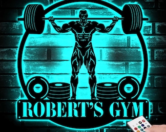 May include: A black metal sign with a silhouette of a weightlifter lifting a barbell. The sign is illuminated with a blue glow and has the text "ROBERT'S GYM" on the bottom.