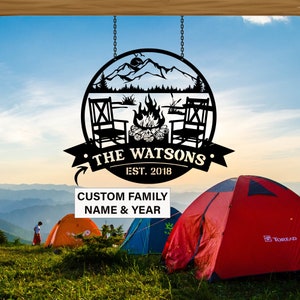 May include: Black metal sign featuring a mountain scene, a campfire, and two chairs. The sign reads "The Watsons Est. 2018". The sign is suspended from a chain and is positioned against a blue sky with white clouds. There are red, orange, and blue tents in the background.