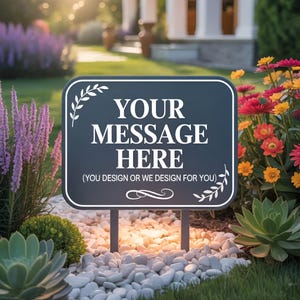 May include: A dark blue rectangular yard sign with white text that reads "YOUR MESSAGE HERE". The sign has a white border and decorative leaf accents. It is mounted on two metal posts and surrounded by white stones and greenery.