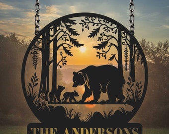 May include: Black metal silhouette of a mama bear and two cubs walking through a forest with a sunset in the background. The text "THE ANDERSONS" is on a separate plaque below the circle.