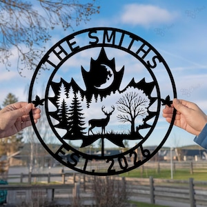 Pode incluir: Placa de nome de família em metal preto com um design de folha de bordo. A placa apresenta um veado, árvores e uma lua. As palavras "THE SMITHS" e "EST. 2026" são exibidas em uma moldura circular.