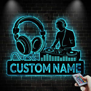 Op de afbeelding: Een zwart en blauw neonbord met een DJ-silhouet met koptelefoon en draaitafels, tegen een bakstenen muur. Het bord bevat de tekst "CUSTOM NAME". Een afstandsbediening is rechtsonder zichtbaar.