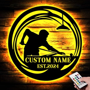May include: A circular black metal wall sign featuring a backlit yellow LED silhouette of a person playing billiards with a hanging lamp above. The sign includes the text CUSTOM NAME and EST.2024 in yellow, accompanied by a remote control for lighting.