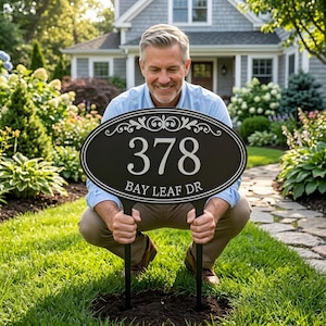 Könnte beinhalten: Ein ovales, schwarzes Adressschild mit der Nummer 378 und dem Straßennamen "Bay Leaf Dr" in weißen Buchstaben. Das Schild wird von einer Person in einem Garten gehalten, mit einem Haus im Hintergrund.