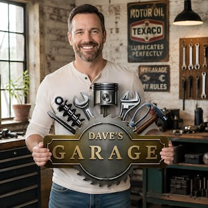 May include: A decorative metal sign featuring the text DAVE'S GARAGE in gold lettering, centered on a circular gear-shaped backing. The sign is adorned with a metallic piston, wrenches, a hammer, and a shock absorber attached to the top edge.
