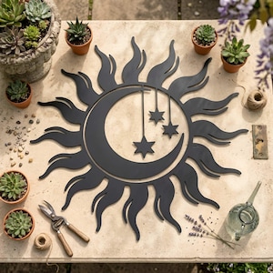 May include: Black metal sun and moon wall art with hanging stars. The celestial design is surrounded by a sunburst pattern. The art is displayed on a stone surface, surrounded by potted succulents, gardening tools, and a watering can.