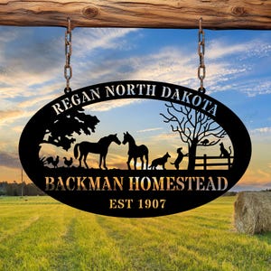 May include: Black metal oval sign with the text "Regan North Dakota" and "Backman Homestead Est 1907". The sign features silhouettes of horses, dogs, a cat, chickens, and a tree, hanging from chains against a sunset.