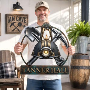 May include: A circular metal wall sign featuring a central miner's helmet with a glowing light, flanked by a crossed shovel and pickaxe, enclosed in a ring. Below the circular frame is a rectangular plaque with the text TANNER HALL in bold letters.