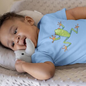 May include: A baby wearing a light blue bodysuit with a green frog graphic. The baby is lying on a white and gray textured surface.