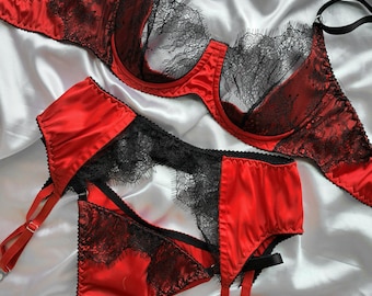 May include: A red and black lingerie set consisting of an underwire bra with black lace detailing and matching high-waisted knickers with integrated garter straps. The set features satin-like fabric, adjustable black straps, and scalloped lace edges.