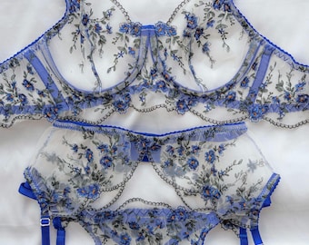 May include: A delicate lingerie set featuring a bra, garter belt, and thong. The set is made of sheer white fabric with blue floral embroidery and blue trim. The bra has adjustable straps and underwire support.
