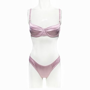 May include: A light pink satin lingerie set, including a bra and matching underwear. The bra has adjustable straps and a gathered design. The underwear is a low-rise style. The set is displayed on a white mannequin against a white background.