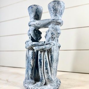 May include: A blue ceramic candle holder featuring three figures holding each other. The figures are stylised and have a rustic, weathered look.