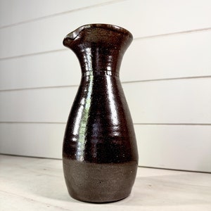 May include: A dark brown ceramic vase with a wide mouth and a slightly flared lip. The vase has a speckled glaze and a smooth, glossy finish.