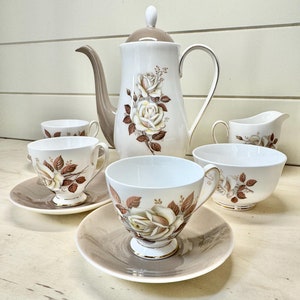 May include: A vintage tea set with a white background and a floral design featuring brown roses. The set includes a teapot, creamer, sugar bowl, and four teacups and saucers.