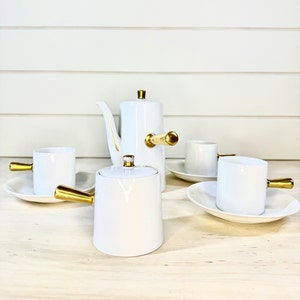 May include: White porcelain tea set with gold accents. The set includes a teapot, creamer, and two cups and saucers. The teapot and creamer have gold handles.