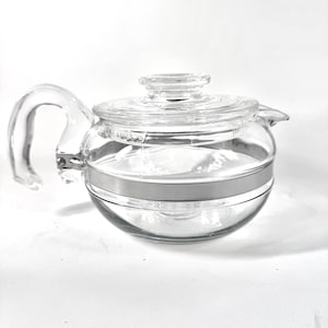 May include: A clear glass teapot with a curved handle and a lid. A silver-coloured metal band encircles the middle of the teapot. The teapot is designed for brewing tea and has a spout for pouring.