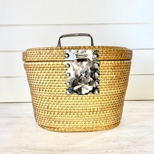 May include: A woven wicker basket with a silver latch and handle. The basket is oval shaped and has a light brown colour.