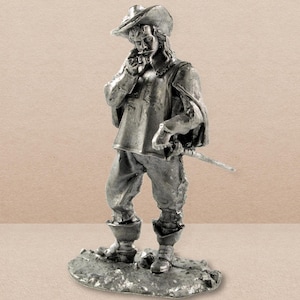 May include: A pewter figurine of a man in a feathered hat and a long coat, holding a sword. The figurine is standing on a base.