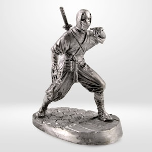 May include: A silver-colored figurine of a ninja in a fighting stance, with a sword on its back. The ninja is wearing a hooded mask and traditional clothing. The figurine is standing on a stone-like base.