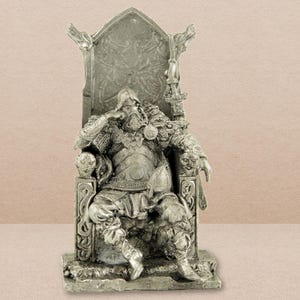 May include: A pewter figurine of a Viking warrior seated on a throne, holding a sword. The warrior is wearing armour and a helmet with horns. The throne is decorated with intricate carvings.