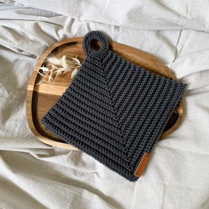 Könnte beinhalten: Ein dunkelgrauer gehäkelter Topflappen mit einer Schlaufe. Der Topflappen liegt auf einem Holztablett mit einem kleinen Zweig getrockneter Blumen.