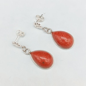 Op de afbeelding: Een paar zilveren oorhangers met druppelvormige, rood-oranje stenen. De stenen zijn gezet in een zilveren rand en hangen aan een bloemvormige oorsteker. De oorbellen worden gepresenteerd tegen een witte achtergrond.