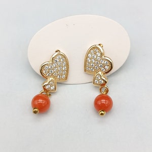 May include: A pair of gold earrings with a dangling orange bead. Each earring features two interlocking heart-shaped charms encrusted with clear cubic zirconia stones.