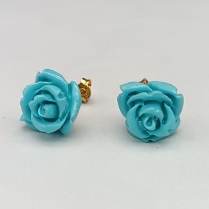 Puede incluir: Un par de pendientes de botón con forma de rosa turquesa con postes dorados. Los pendientes son de un tono azul vibrante y están colocados sobre un fondo blanco liso. Los pétalos de rosa son detallados y esculpidos.