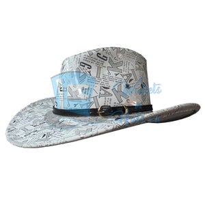 Peut inclure: Un chapeau de cowboy blanc orné d'un imprimé noir et gris composé de diverses lettres et extraits de texte. Le chapeau comprend une fine bande noire autour de la calotte avec une petite boucle en métal doré et deux œillets métalliques argentés.
