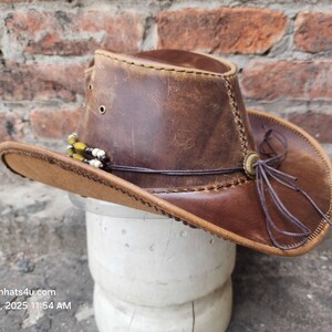 May include: A brown leather cowboy hat with a brown leather band and a brown leather strap. The hat has a distressed look and is decorated with a beaded trim.