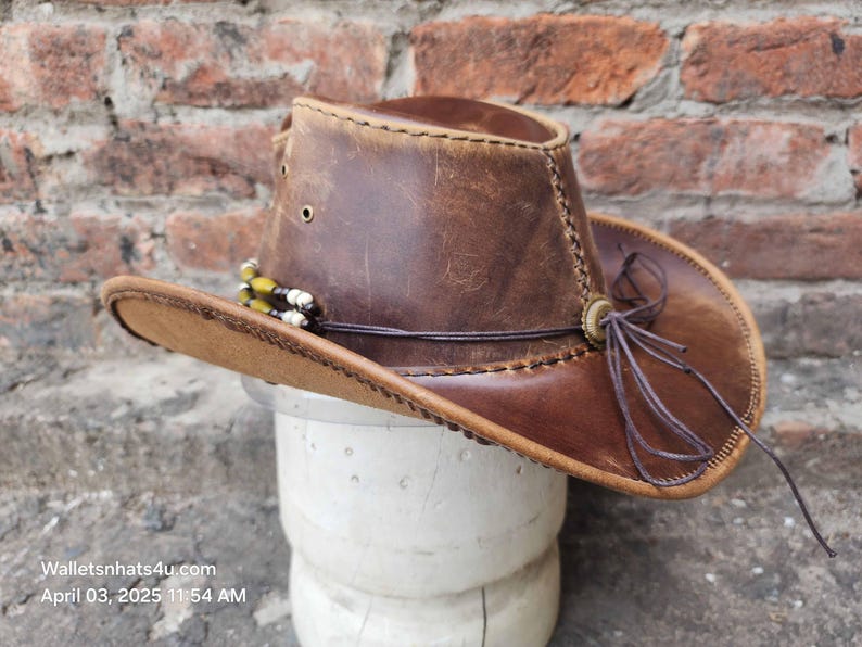 May include: A brown leather cowboy hat with a brown leather band and a brown leather strap. The hat has a distressed look and is decorated with a beaded trim.