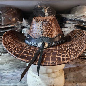 May include: A brown leather cowboy hat with a crocodile pattern. The hat has a black band with a silver buckle and a black braided trim.
