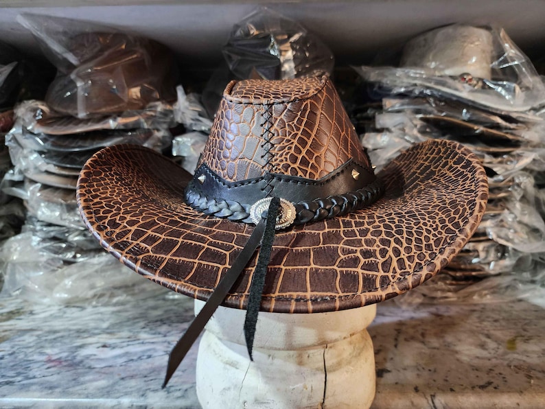 May include: A brown leather cowboy hat with a crocodile pattern. The hat has a black band with a silver buckle and a black braided trim.