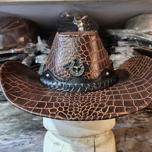 May include: A brown leather cowboy hat with a crocodile pattern and a silver horseshoe design. The hat has a black band with a braided design.