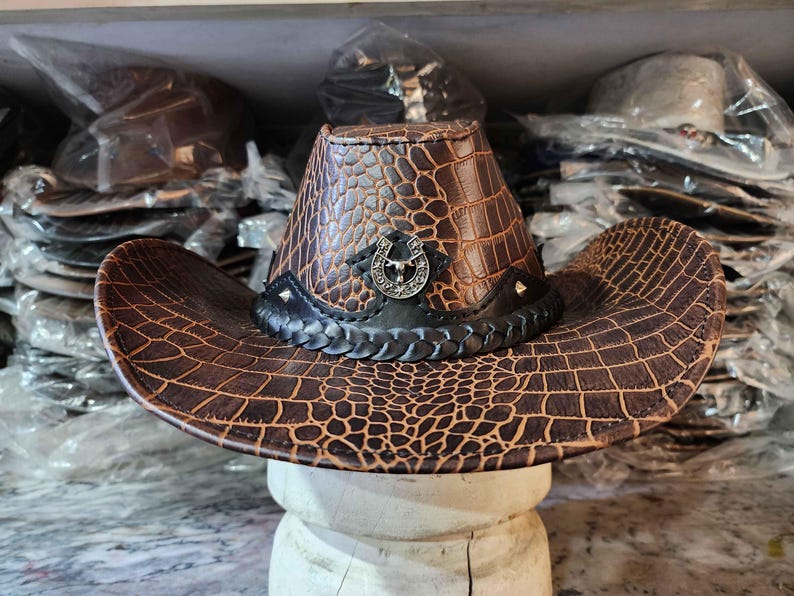 May include: A brown leather cowboy hat with a crocodile pattern and a silver horseshoe design. The hat has a black band with a braided design.