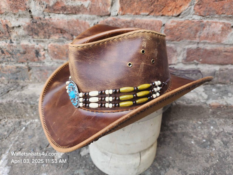 May include: A brown leather cowboy hat with a turquoise and silver concho and a beaded band with white and green beads.