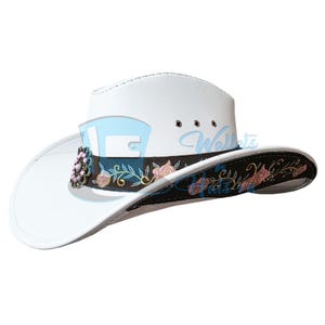 Puede incluir: Sombrero de vaquero blanco con una banda decorativa con bordados florales en rosa y azul. El sombrero tiene un ala curvada y una banda decorativa con bordados florales rosas y azules.