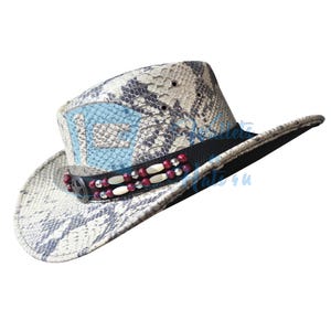 May include: Cowboy hat with a snakeskin pattern in shades of gray, white, and blue. The hat features a black band with decorative red and white beads and a star-shaped embellishment. The brim is curved, and the crown has a light blue design.