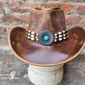 May include: A brown leather cowboy hat with a turquoise and silver concho and a white beaded band.